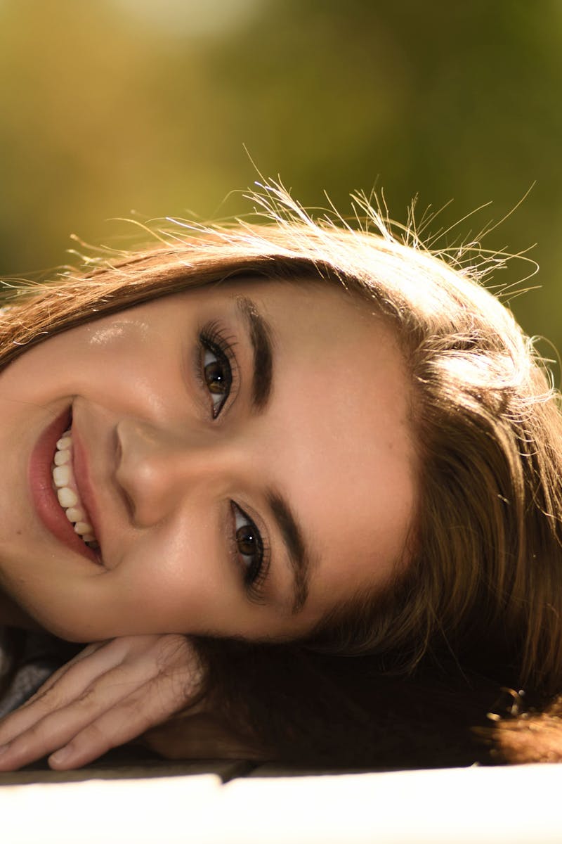 A young woman with long hair and a radiant smile lies on the grass during a sunny day.